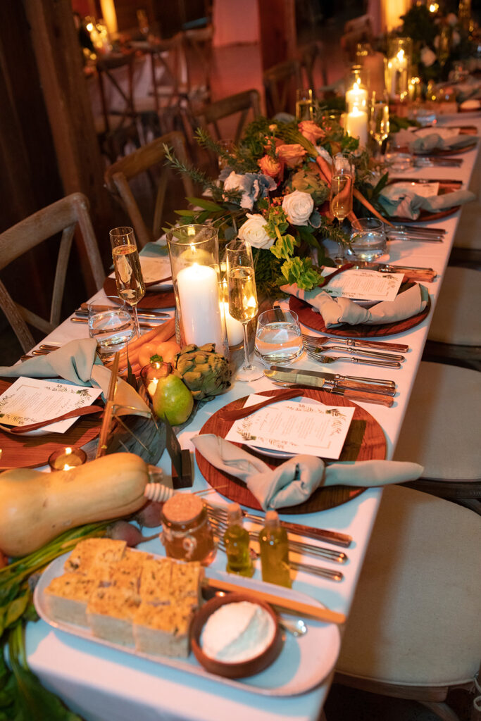 reception table set with wood chargers, modern brushed gold flatware, and a garland of warm lit pillar candles and fresh green and orange produce