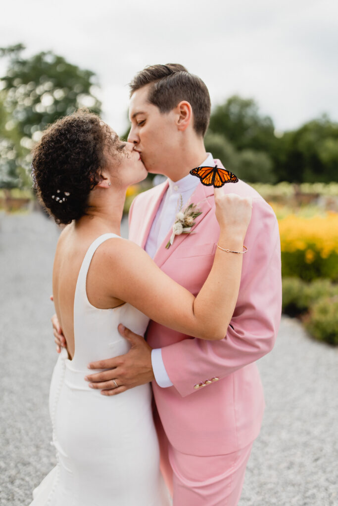 bride in blush suit and bride in white dress kiss outside with monarch butterfly resting on hand