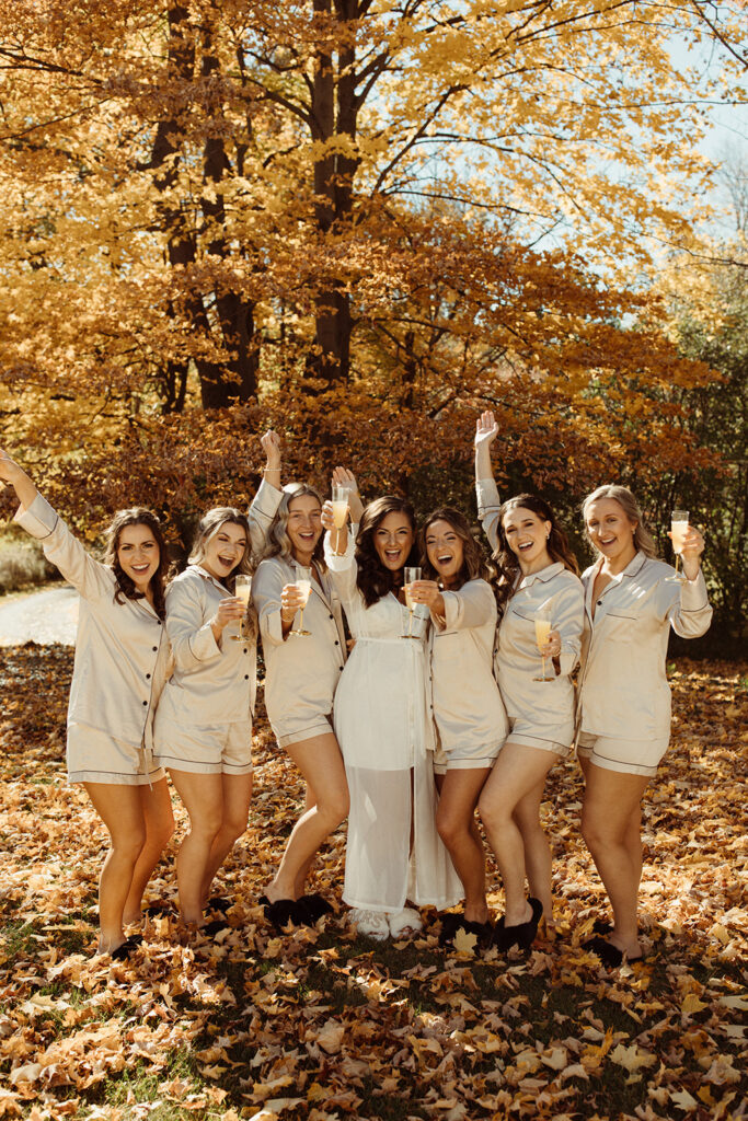 bridesmaids in blush matching pajamas and bride raise glasses outside in front of fall foliage