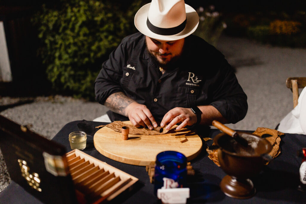 man in black shirt and fedora hand rolls cigars for wedding guests