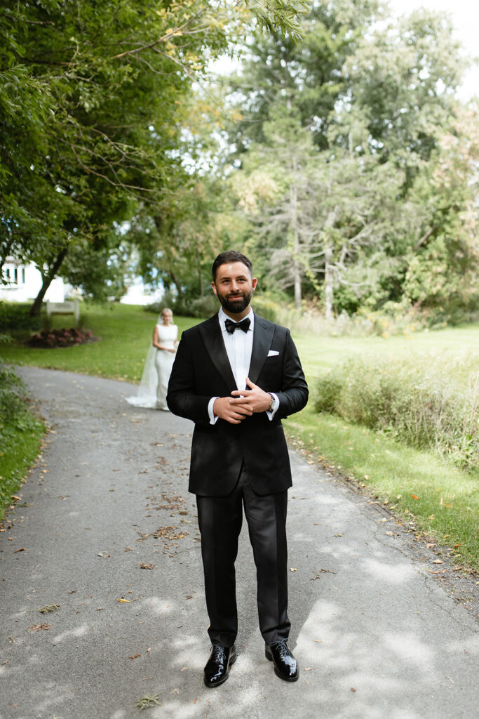 groom waiting on a tree lines path for bride to greet him for their outdoor first look in Upstate New York