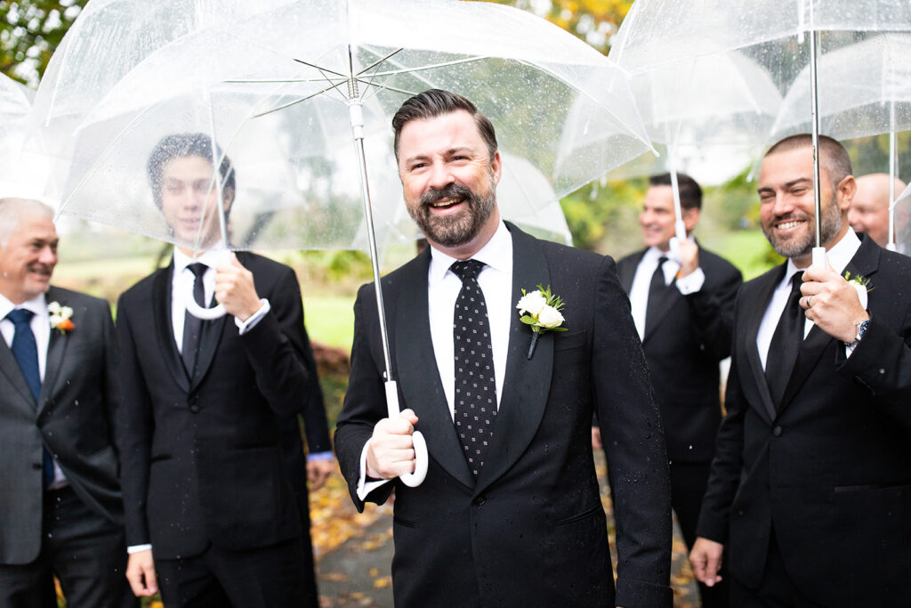 Groom walking in the light rain with groomsmen and clear bubble umbrellas 