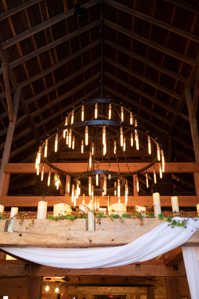 large edison chandelier in vaulted barn ceiling and a beam lines with pillar candles and greenery  