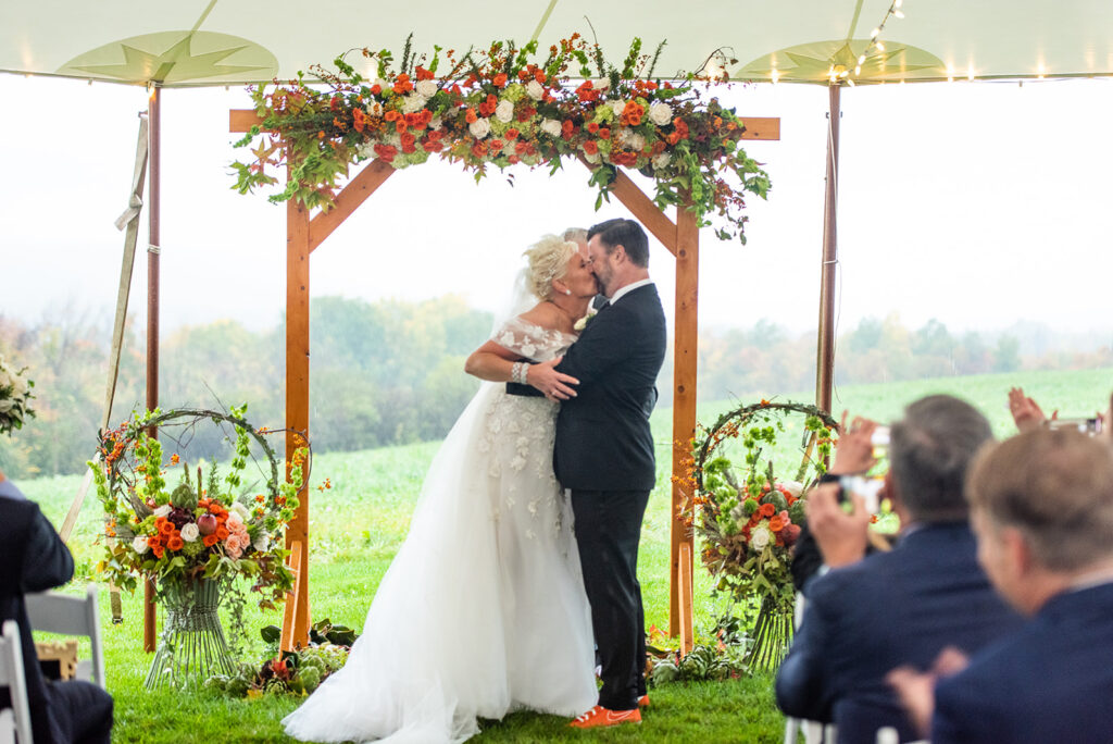 bride kissing groom under a colorful orange and green wedding arbor in a sailcloth tent upstate