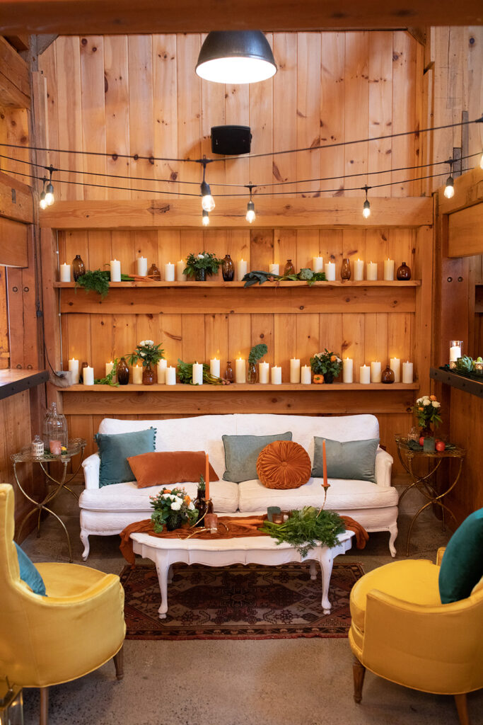 white, gold, teal, and orange vintage lounge tucked into a nook in a barn wedding venue with pillar candles on the wall behind