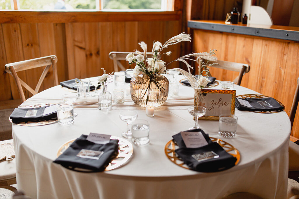round table ready for guests with white table cloth, back napkins, copper chargers and neutral flowers