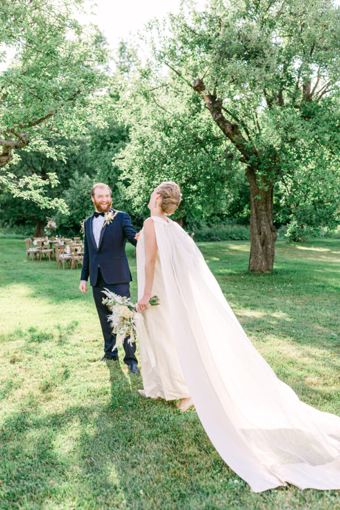 bride with flowing white cape and groom walk outside in the summer sun