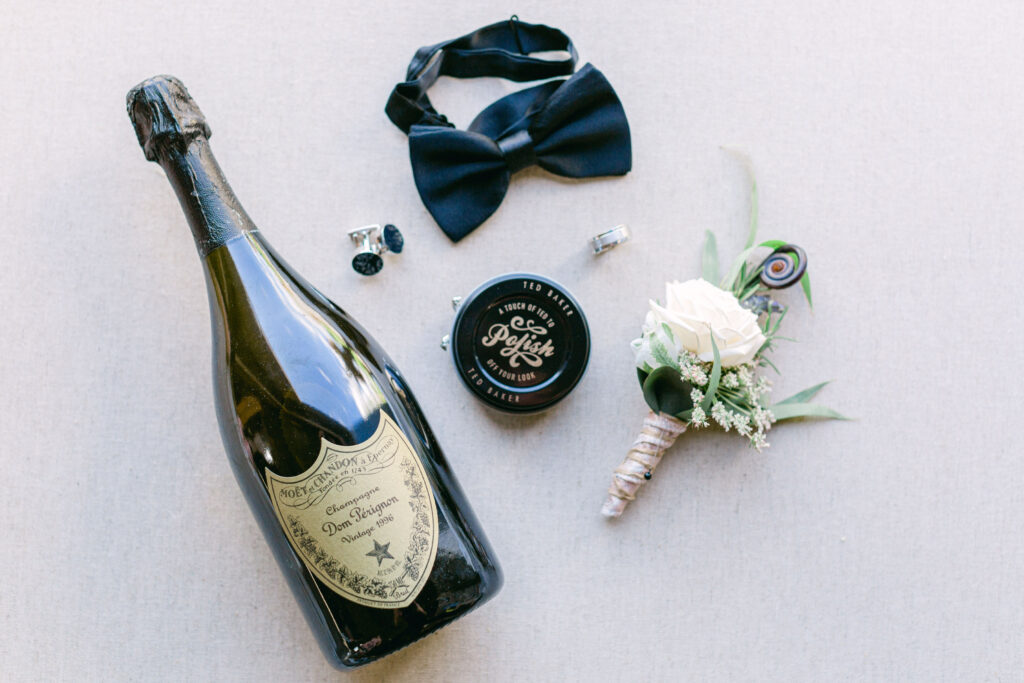 a flatlay against natural linen with black bow tie, white floral boutonniere, cufflinks and wedding band and a bottle of french champagne 
