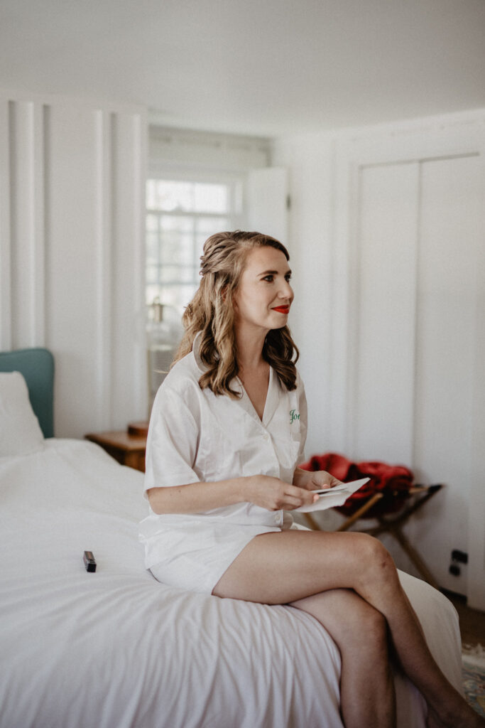 bride in white pajamas sits on white sheets of a bed in the farmer cottage of weekend wedding venue in upstate new york